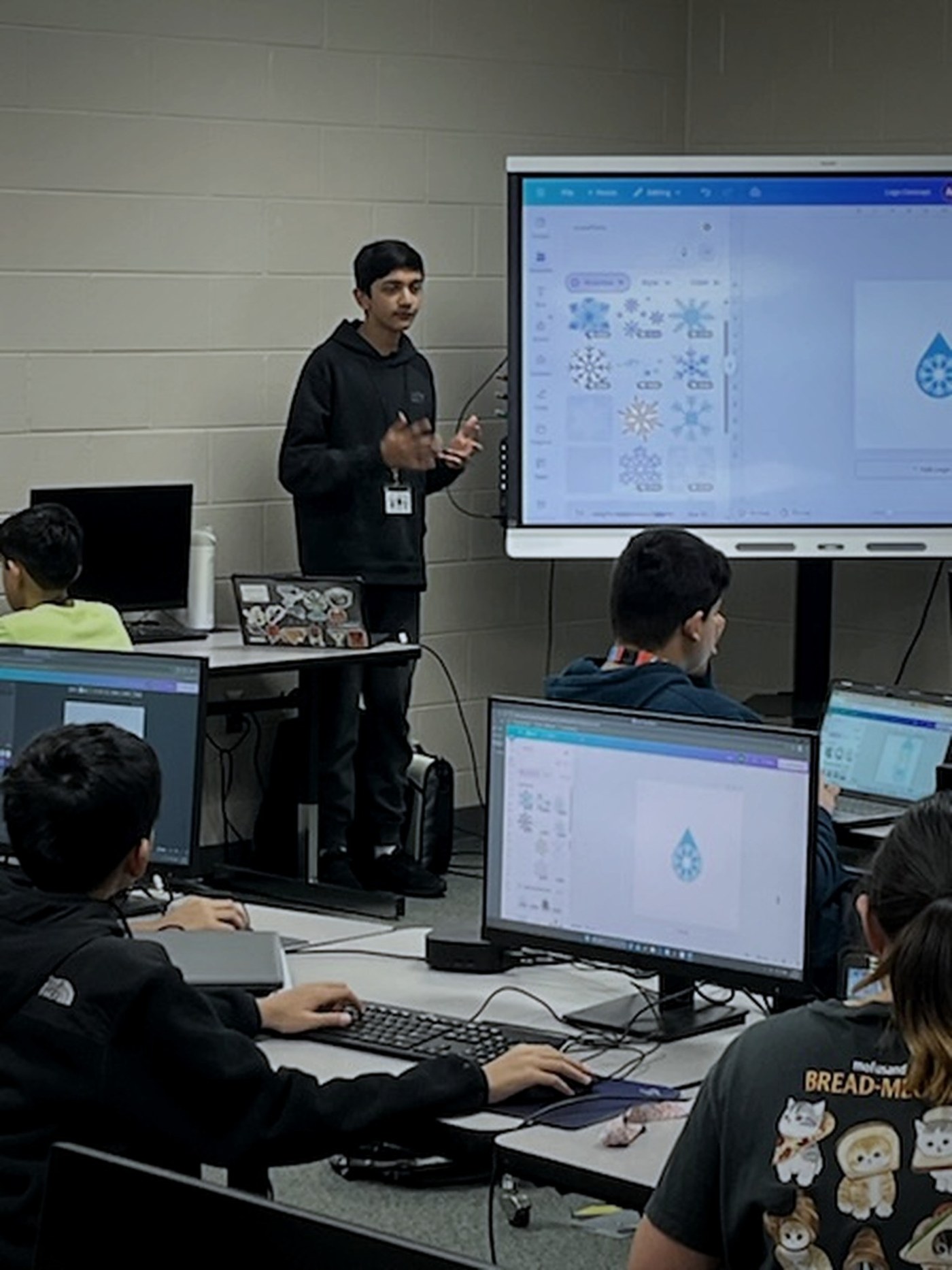Aariv at a classroom display explaining a logo in Canva while members work at computers.